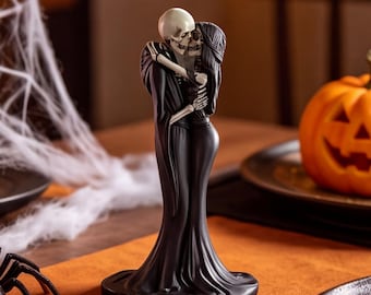 May include: A decorative figurine featuring two skeletons in a close embrace, both wearing long, flowing black robes. The skeletons are positioned face-to-face, with their arms wrapped around each other in a standing pose.