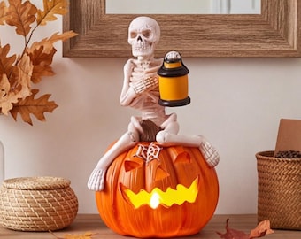 Puede incluir: Una figura de esqueleto blanco sentada sobre una calabaza naranja tallada. El esqueleto sostiene una pequeña linterna negra y amarilla en una mano. La calabaza presenta una boca dentada brillante y un diseño de telaraña blanca.