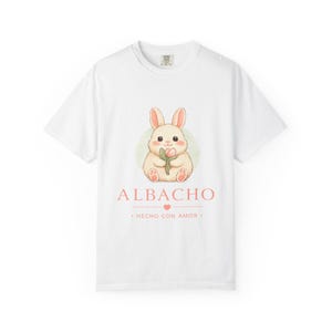 Peut inclure: Un t-shirt blanc à manches courtes avec un motif représentant un lapin brun clair tenant une petite fleur rose. Sous le lapin, le texte indique ALBACHO, suivi d'une petite icône de cœur et du texte HECHO CON AMOR en rose clair.