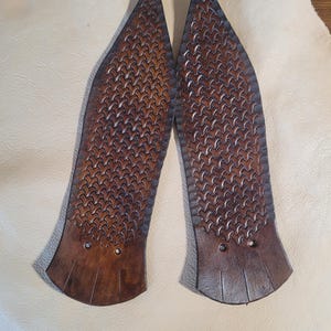 May include: Two elongated, pointed leather boot tips in a rich brown tone, featuring a detailed basket-weave embossed pattern across the surface. Each piece has a smooth, solid base with two small circular holes and three vertical cut lines.