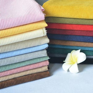 Puede incluir: Una pila de tela de pana doblada en varios colores, incluyendo rosa, amarillo, crema, azul claro, gris, verde y marrón, junto a una pila de tonos más oscuros como mostaza, oliva, rojo, azul marino y negro, con una flor de plumeria blanca.