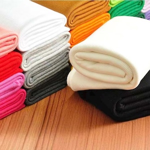 Puede incluir: Una colección de piezas de tela acanalada dobladas en una variedad de colores sólidos que incluyen blanco, negro, rojo, rosa, naranja, amarillo, verde y morado. Cada pieza de tela presenta una textura acanalada vertical en todo el material.