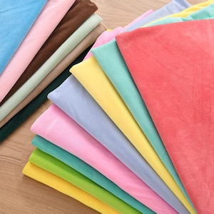 Può includere: Una collezione di pezzi di tessuto piegati in una varietà di colori solidi tra cui corallo, giallo, verde chiaro, rosa, azzurro, menta e marrone. I tessuti sono impilati in strati sovrapposti, mostrando una superficie liscia e morbida.
