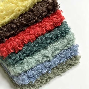 Puede incluir: Una pila de siete muestras de tela dobladas con una superficie de pelo rizado y texturizado. Los colores de arriba a abajo incluyen amarillo, marrón oscuro, rojo brillante, verde azulado oscuro, verde salvia claro, azul bígaro y verde oliva.