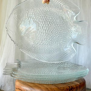Op de afbeelding: Een set doorzichtige glazen borden in de vorm van een vis, met een gestructureerd schubbenpatroon op het lichaam en lineaire details op de vinnen. De borden zijn transparant met een glanzende afwerking en tonen een duidelijk vissilhouet.