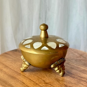 May include: A small, round brass lidded container featuring three ornate, textured legs and a central knob handle. The lid is decorated with a series of inlaid, white, teardrop-shaped mother-of-pearl accents arranged in a circular pattern.