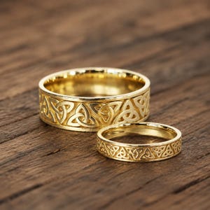 Könnte beinhalten: Zwei goldene Ringe mit einem durchgehenden, geprägten Triquetra- oder Dreiecks-Knotenmuster auf dem äußeren Band. Der größere Ring ist breiter als der kleinere, und beide weisen eine polierte metallische Oberfläche mit komplizierten, sich wiederholenden Knotenmustern auf.