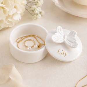 Puede incluir: Un joyero de cerámica blanca con tapa a juego que presenta un asa esculpida en forma de mariposa. La tapa está personalizada con el texto dorado Lily. Dentro del recipiente hay un collar de cadena dorada con colgante de perla y un anillo de perla.