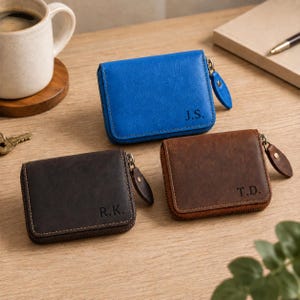 May include: Three leather zippered wallets in blue, dark brown, and medium brown. Each wallet features a metal zipper with a matching leather pull tab and is personalized with embossed initials J.S., R.K., and T.D. on the front.