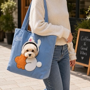 Pode incluir: Uma bolsa de transporte para animais de estimação em lona azul-clara com uma abertura circular para um cão pequeno espreitar. A bolsa é decorada com um aplique de coelho branco e laranja e o nome Bill bordado em branco na lateral.