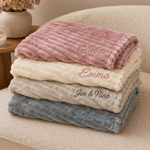 May include: A stack of four folded plush blankets with a ribbed texture. The top blanket is dusty rose with the name Olivia embroidered. The second is cream with Emma embroidered. The third is beige with Jim & Nina. The bottom is grey with The Smiths.