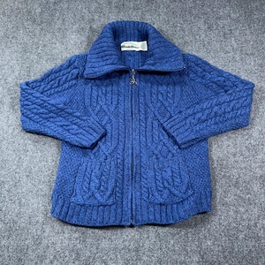 May include: A royal blue cable-knit zip-up sweater featuring a high collar, long sleeves, and two front pockets. The garment has a full-length metal zipper with a decorative pull tab and a white rectangular brand label sewn inside the neckline.