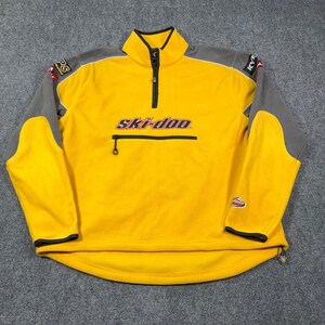 May include: A yellow and gray quarter-zip fleece pullover featuring the Ski-Doo logo on the chest. The garment includes a front zippered pocket, a high collar, and black trim on the cuffs and hem. Various brand logos are visible on the shoulders.