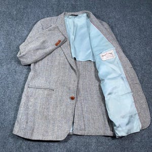 May include: A grey herringbone blazer featuring a light blue interior lining, a single brown button closure on the front, and three brown buttons on each sleeve cuff. A white Harris Tweed label is stitched onto the interior lining of the garment.