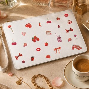 Può includere: Una pochette rettangolare bianca con cinturino da polso, caratterizzata da un motivo ripetuto di piccole illustrazioni rosse tra cui fragole, ciliegie, labbra, fiocchi, cuori, una coccinella, un peperoncino, un bicchiere di succo e vari dolci.