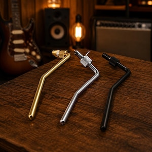 Könnte beinhalten: Drei Gitarren-Tremolo-Arme in Gold, Silber und Schwarz. Jeder Metallarm hat ein gebogenes, abgewinkeltes Design mit einem Gewindeende und einem geriffelten Einstellknopf. Die Komponenten sind nebeneinander auf einer Holzoberfläche angeordnet.