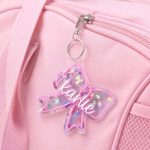 May include: A clear acrylic bow-shaped keychain with a pink outline, filled with colorful butterfly-shaped glitter, featuring the name Karlie in white script lettering, attached to a silver metal keyring on a light pink fabric bag.