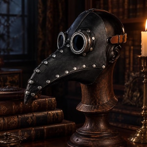 Op de afbeelding: Een zwart pestmeestermasker met een lange, puntige snavel, ronde ooglenzen met metalen randen en zilverkleurige klinknagels langs de naden. Het masker is bevestigd met een bruine leren riem en gesp en staat op een donkere houten standaard.