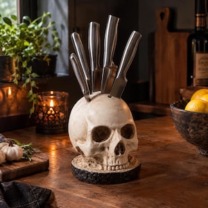 Può includere: Un ceppo portacoltelli a forma di teschio umano realizzato in materiale color osso, dotato di cinque fessure sulla parte superiore che ospitano cinque coltelli da cucina in acciaio inossidabile con manici in metallo su una base circolare scura.