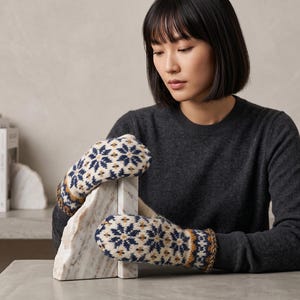 May include: A pair of knit mittens featuring a cream-colored base with a repeating navy blue and mustard yellow snowflake pattern. The mittens have a ribbed cuff and a detailed geometric border design around the wrist area, showcasing intricate stitching.