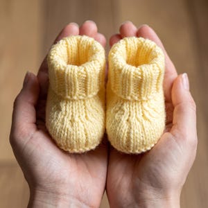 以下が含まれることがあります： リブ編みのカフと前面にテクスチャーのあるケーブル編み模様が特徴の、淡い黄色のニット製ベビー用ブーティ一足。柔らかい毛糸で作られており、丸みを帯びた爪先と、編み目がはっきりと見えるクラシックな構造が特徴です。