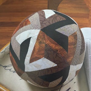May include: A spherical fabric pincushion featuring a geometric patchwork design. The surface is composed of triangular and diamond-shaped fabric segments in shades of black, white, cream, and various brown tones, some with subtle leaf or dot patterns.