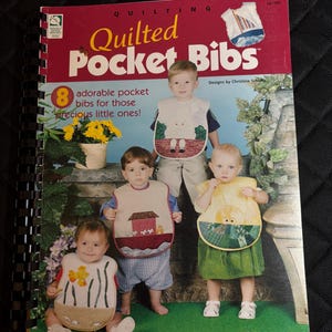 Könnte beinhalten: Ein spiralgebundenes Anleitungsbuch mit dem Titel Quilted Pocket Bibs von Christine Schultz. Das Cover zeigt ein Foto von vier Kindern, die verschiedene gesteppte Lätzchen mit Motiven wie einem Schaf, einem Haus, einer Sonne und einem Fisch tragen.