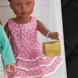 Puede incluir: Una muñeca de piel oscura y cabello oscuro peinado en dos moños, viste un vestido rosa sin mangas con un patrón circular blanco y dos niveles de volantes. La muñeca sostiene un pequeño bolso dorado rectangular con asa de cuentas. El texto D,G es visible.
