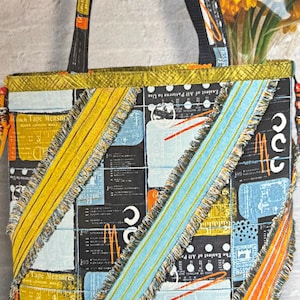 May include: A fabric tote bag featuring a patchwork design of black, yellow, and light blue panels with sewing-themed graphics and text. The bag has two black straps and decorative diagonal fabric strips with frayed edges in yellow and blue.
