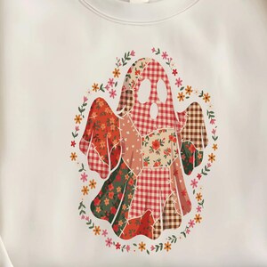 May include: A cream-coloured crewneck sweatshirt featuring a central graphic of a ghost composed of various patchwork patterns, including red and white gingham, floral prints, and brown plaid, surrounded by a wreath of small colourful flowers.