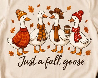 Puede incluir: Una camiseta de color crema con un gráfico de cuatro gansos que llevan accesorios de otoño como bufandas, gorros y jerséis, con hojas cayendo y el texto "Just a fall goose" impreso en una fuente cursiva oscura bajo la ilustración.
