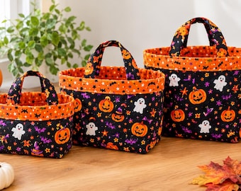 Puede incluir: Tres cestas de tela de tamaños graduados con fondo negro y un colorido patrón de Halloween de calabazas naranjas, fantasmas blancos, murciélagos morados y estrellas. Cada cesta tiene dos asas y un borde de color naranja.