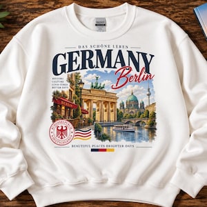Può includere: Una felpa bianca a girocollo con una stampa grafica di Berlino, Germania. Il design include il testo "DAS SCHÖNE LEBEN GERMANY Berlin", la Porta di Brandeburgo, il Duomo di Berlino, un battello fluviale, la bandiera tedesca e un'aquila.