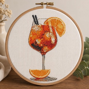 May include: A cross-stitch embroidery in a round wooden hoop featuring a vibrant orange cocktail in a stemmed glass. The drink is garnished with an orange slice on the rim and another slice at the base, complete with two black straws.