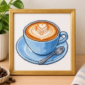 Könnte beinhalten: Ein gerahmtes Kreuzstich-Stickbild mit einer hellblauen Kaffeetasse und Untertasse auf weißem Grund. Die Tasse enthält einen Latte mit einem weißen, herzförmigen Milchschaummuster und einem kleinen silbernen Löffel, der auf der Untertasse liegt.