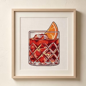 Peut inclure: Une broderie au point de croix encadrée représentant un verre à whisky rempli d'un liquide ambré, de glaçons et d'une tranche d'orange. Le verre présente un motif en losanges texturé et est centré dans un cadre en bois clair.