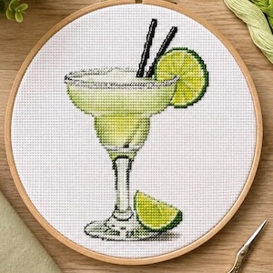 Puede incluir: Un proyecto de bordado en punto de cruz en un bastidor de madera redondo que muestra un cóctel margarita en una copa de tallo. La bebida es de color verde claro con borde salado, dos pajitas negras y una rodaja de lima como adorno.