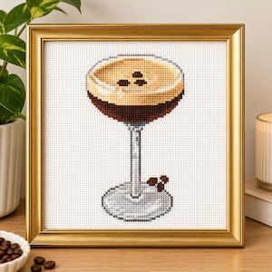 Peut inclure: Une broderie au point de croix dans un cadre doré représentant un espresso martini dans un verre à pied. La boisson présente un liquide brun foncé surmonté d'une mousse beige clair et de trois grains de café. Deux grains sont à la base.