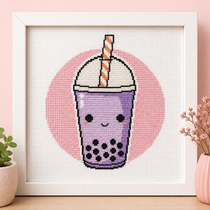 Puede incluir: Un bordado de punto de cruz en un marco blanco que presenta un vaso de té de burbujas morado con una cara sonriente, perlas de tapioca negras y una pajita a rayas naranjas y blancas sobre un fondo circular de color rosa.