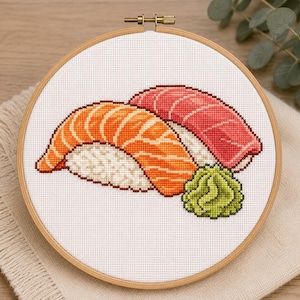 May include: A cross-stitch embroidery project in a round wooden hoop featuring two pieces of sushi, one with orange salmon and one with red tuna, alongside a small mound of green wasabi, all stitched onto a white fabric background.