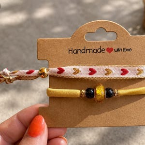 Op de afbeelding: Twee armbanden op een bruine kartonnen displaykaart met de tekst Handmade with love. De bovenste armband is een lichtroze stoffen band met rode en gouden hartjespatronen. De onderste armband is een gele elastische band met twee zwarte kralen en een centrale goudgespikkelde barnsteenkleurige kraal.
