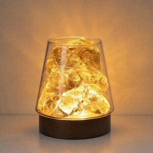 May include: A table lamp featuring a clear, tapered glass shade filled with textured, translucent amber-coloured crystalline shapes. The lamp is supported by a circular, dark brown wooden base that emits a warm, soft glow from within.