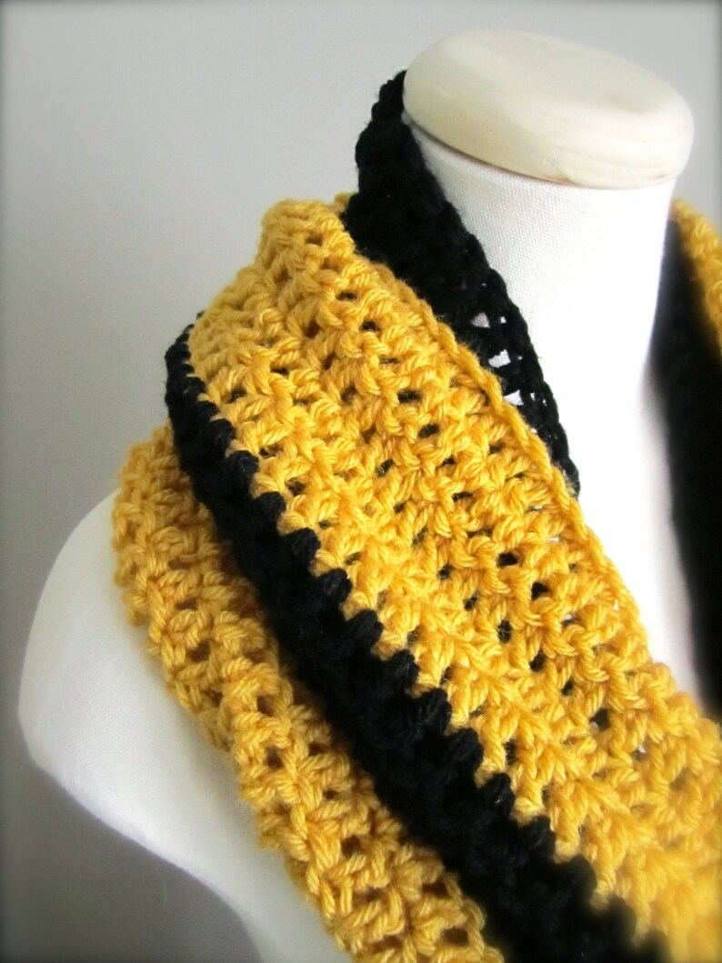 May include: A yellow crocheted infinity scarf with a black crocheted trim. The scarf is displayed on a white mannequin.