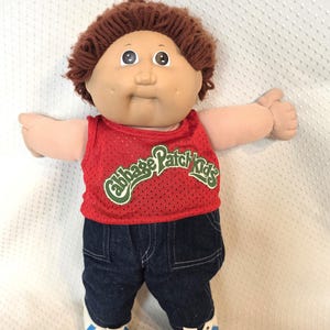 May include: A Cabbage Patch Kids doll with brown yarn hair, a round face, and dimples. The doll wears a red mesh tank top with the green Cabbage Patch Kids logo printed on the front, dark blue denim pants, and white sneakers with blue stripes.