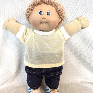 May include: A soft-sculpture doll with tan skin, blue eyes, and light brown yarn hair. The doll wears a short-sleeved yellow and white striped shirt with a white collar, dark blue denim-style pants, and white sneakers with blue stripes.