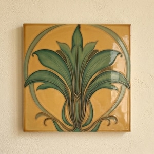 Puede incluir: Un azulejo de cerámica cuadrado con acabado brillante y fondo amarillo mostaza. Un motivo botánico verde estilizado está centrado dentro de un marco circular, mostrando formas de hojas simétricas con contornos y sombreados en verde oscuro.