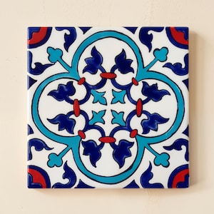 May include: A square ceramic tile featuring a white background with a symmetrical floral pattern in shades of dark blue, bright turquoise, and red. The design consists of a central motif surrounded by an oval border and corner accents.