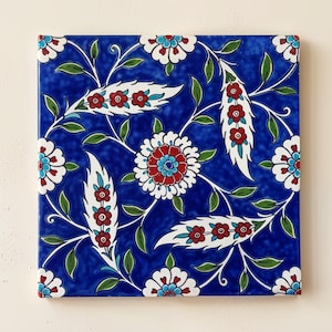 May include: A square ceramic tile featuring a vibrant cobalt blue background with a repeating pattern of white, red, and turquoise floral motifs. The design includes stylized blossoms and elongated, curved leaf shapes with small red flowers.