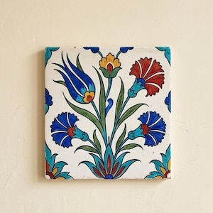 May include: A square ceramic tile featuring a hand-painted floral pattern on a white background. The design includes a central blue tulip, a red carnation, and smaller blue flowers with green stems and leaves, framed by a decorative border.