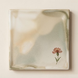 May include: A square ceramic plate featuring a smooth, glazed surface with blended washes of soft sage green, cream, and warm beige tones. A single, detailed illustration of a small, pinkish-orange flower with green leaves is positioned in the lower right corner.